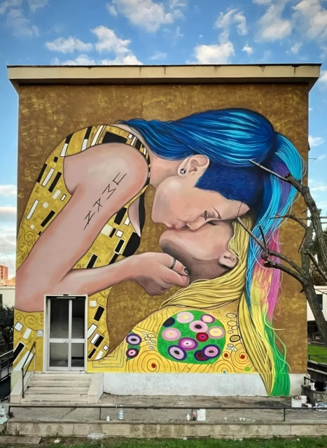 Streetartwall. A modern homage to the painter Gustav Klimt was sprayed/painted on the outside wall of a square, two-storey building. In contrast to the original painting, in which a man is kissing a woman, here two women are kissing. On the left, a young woman with short blue hair is kissing a young woman on the right. She has long blonde hair with pink and green highlights. The couple is depicted from the side. The golden background and clothing are based on the original. They are decorated with black and white rectangular ornaments on the left and colorful circles on the right, both on gold. (In front of the mural there is a stone staircase to the front door on the left and a bare tree on the right).