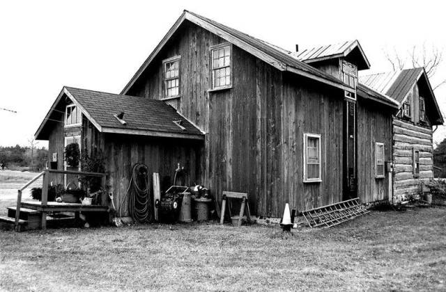 Una casa antigua, grande, de madera, con tejados en dos aguas y buhardilla. Aperos en el exterior. Imagen en blanco y negro.