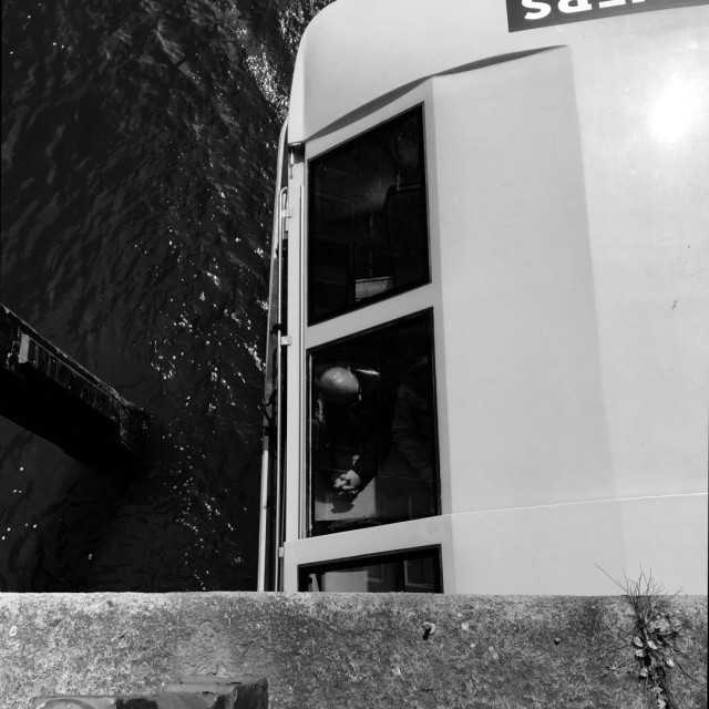 Black and white photo. Looking down from a small bridge onto the roof of a tour boot in Amsterdam, disappearing under the bridge. The roof, seen on the right of the frame,  appears as light grey in this photo. On the left rippling water is visible. In the middle, two windows.