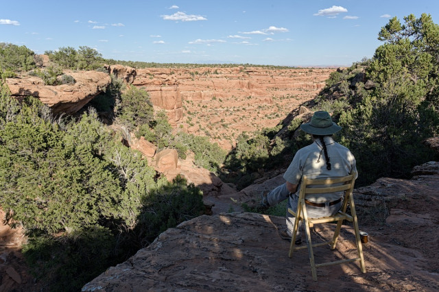 A man sitting in a folding wooden chair on the edge of a large red rock canyon. On the ground to the left is a coffee mug. The man wears a tan shirt, grey pants and a sage green flop hat. A braided ponytail hangs down his back.