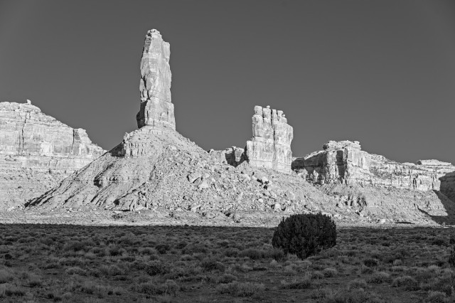 A black and white photo of highly eroded sandstone formations. In a field of sagebrush in the foreground is a round bush. In the background are two tall thin fins sitting atop conical bases of eroded rock.The fin in front at center left is narrower than the fin further back at center right. The background is shear cliffs with  flat tops.