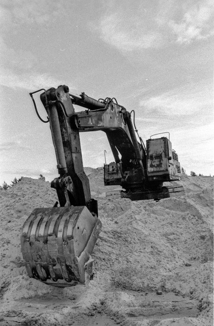 The image is a black and white photo of a Crawler Excavator sitting on top of a pile of sand. The Crawler Excavators is a large machine with a metal scoop at the front. The Excavator is sitting on a large pile of sand that is about twice as tall as the Excavator