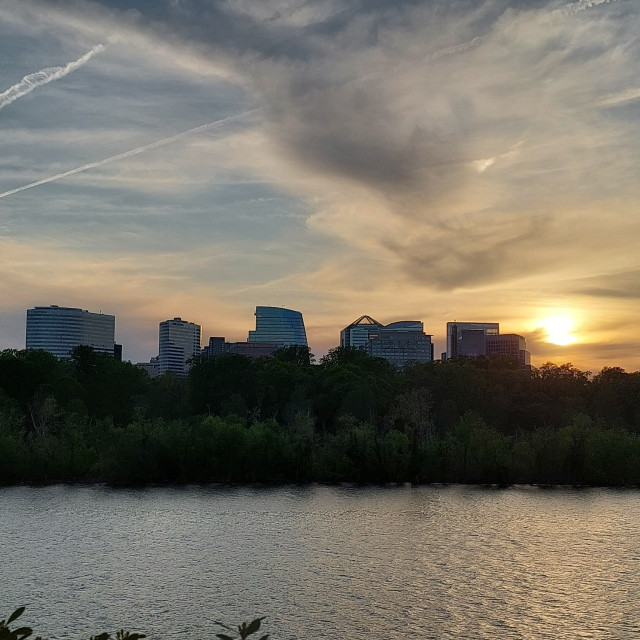 A sunset photo. a somewhat cloudy sky with jet contrails. There are large office buildings in the distance across a river. There are trees between the river's edge and the buildings.