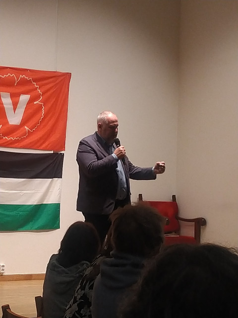Jonas Sjöstedt talar framför en flagga med Vänsterpartiets logga och en palestinsk flagga.