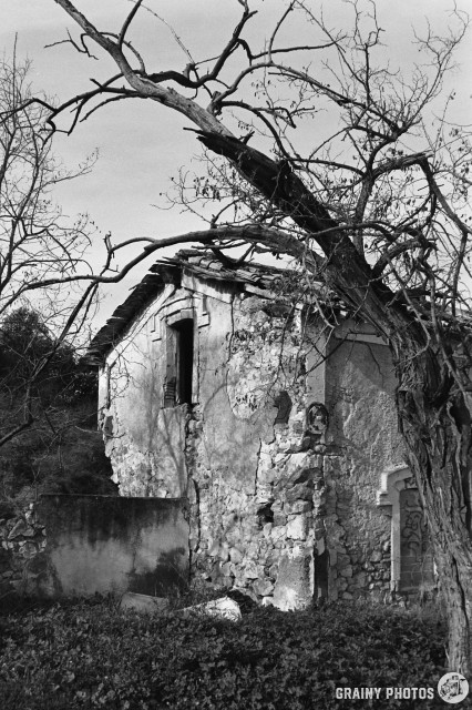 A black-and-white film photo of the gable wall of an abandoned railway worker's cottage. The cottage is amongst trees and is showing signs of decay.