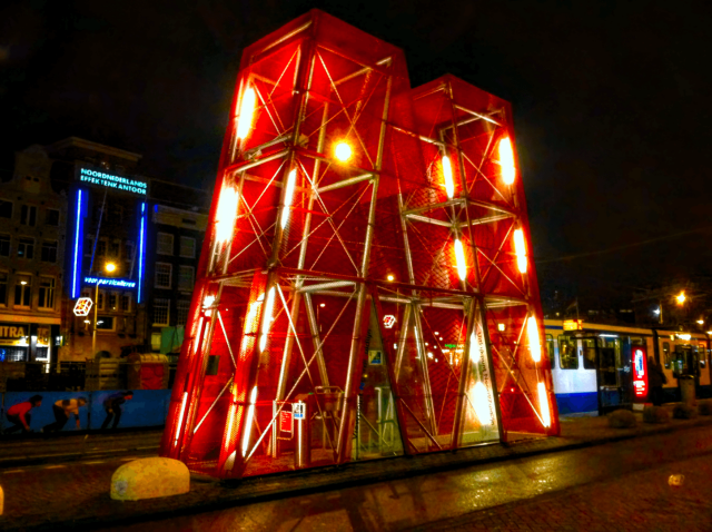 Colour night photo of a large red structure in the shape of a capital letter "M", lit from the inside by several white lights. The structure is on a local street. A blue and white tram can be seen at the right of the frame.