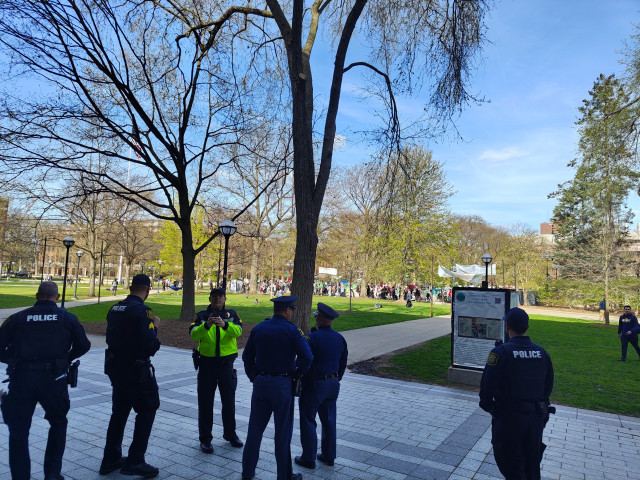 A group of Ann Arbor and Michigan State police stand, watching and surveilling the encampment across the diag. One police officer has his phone out pointed toward the camera, presumably taking a picture or video of the photographer (me).