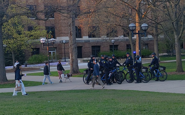 Group of 7 police officers (agency unidentified, likely Ann Arbor or State) stand, mounted on their bikes, watching the encampment. Students walk by.