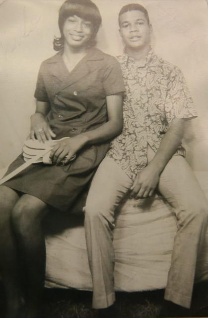 Sepia-toned photo of a couple in a casual seated pose, looking towards the camera. She is on the left, nearly sitting on his lap, smiling, wearing a dark knee-length dress with buttons up the front and a wide collar, holding a white purse. Her hair is straight, shoulder-length, curled at the end, with bangs. He is wearing light-colored slacks and a boldly patterned shirt, like a Hawaiian shirt, and has his arm around his wife. His hair is short and his expression is more curious than friendly.