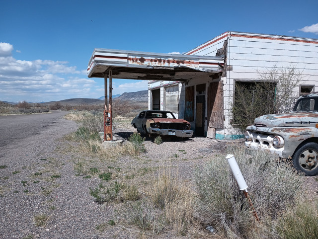 Abandoned gas station. To the right is the front of a '50s era truck. A car sits on blocks under the gas pump Island canopy. The front door and windows are boarded up. An abandoned road runs off to the left of the frame.