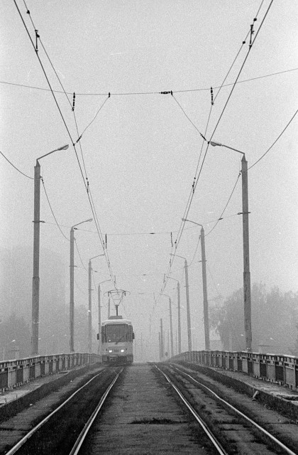 The image shows a tram emerging from a thick fog as it crosses a bridge. The tram is moving toward the observer. There are two tracks. There are pillars along the edges of the bridge. The pillars, taut wires and rails create a perspective moving away from the observer into the fog.
The fog is a thick, white cloud that obscures most of the surroundings. The fog is so dense that it is difficult to see more than a few feet behind the tram. The fog creates an eerie and mysterious atmosphere.
