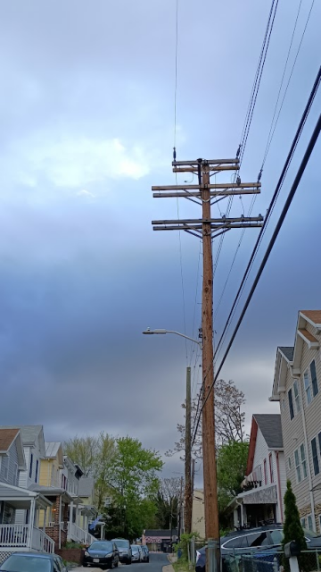 Big grey clouds with a patch of blue obscure the morning sun, looming over some trees, parked cars, a side road, row houses, and a utility pole and power wires in the foreground. It's 7:30am, cool and still slightly quiet.