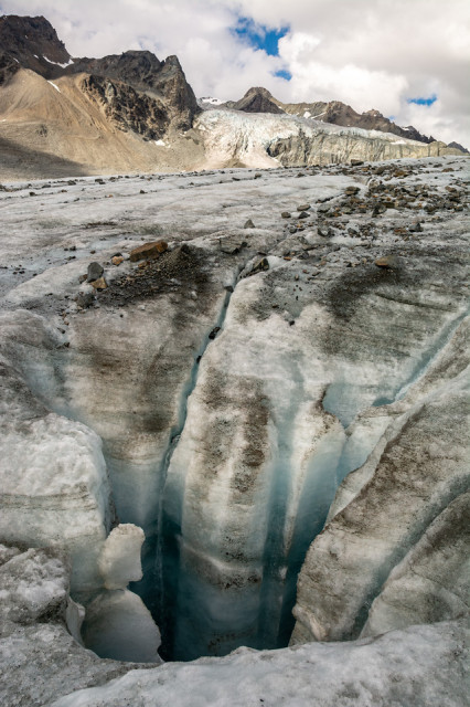 The photo presents a dramatic vertical view over a deep moulin on the Gulkana Glacier. The moulin, a cylindrical, well-like ice feature formed by running water, creates a stark contrast of deep azure within the surrounding white and gray glacial surface. Jagged rocks and gravel are scattered across the ice, leading up to the impressive Gabriel Icefall, which stands out in the distance against the rugged peaks of the Eastern Alaska Range. Patches of blue sky are visible above, with sunlight accentuating the icefall's textures and contours.