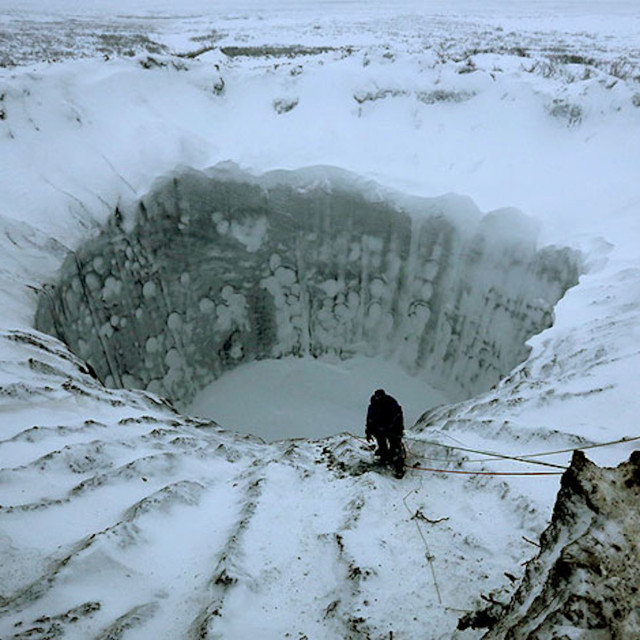 Photo of a wide and deep hole in the permafrost above the Arctic Circle, with a person standing to the bottom of the pit. 