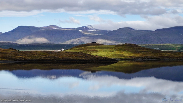 A photo of a summer landscape. The foreground is a body of almost still water, in the middle of the shot is green and brown vegetation across a small hook of land. A few small buildings can be seen, and a dozen or so white dots which are either swans or geese. In the background is a range of bare, brown-grey mountains, with dappled sunlight across the flanks. A little snow is visible as stripes near the peaks. Above them is blue sky with a layer of woolly grey cloud above. The whole scene is reflected in the still waters of the lake.