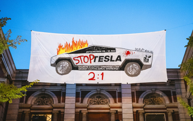 "Stop Tesla" steht auf einem Transparent an der Roten Flora. Der abgebildete Cybertruck brennt. Das Plakat ist ein Aufruf für die Aktionstage von Dirupt. Unter der Abbildung steht in rot: 2:1