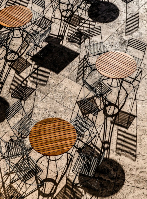 Photography. A color photograph
of about 12 bistro chairs and three tables. The view is from above of bistro furniture in the sunlight. The chairs have the typical striped look, with the struts of the backrest and seat also creating a striped shadow pattern on the floor. Together with the round wooden tables, which are also striped, the result is a chaotic picture of stripes, shadows and circles. In addition, there are square beige-colored stone slabs, whereby the interplay of light, shadow and substance may initially appear confusing to the viewer, but on closer inspection reveal a clear order.
Info: Camera Nikon D800E