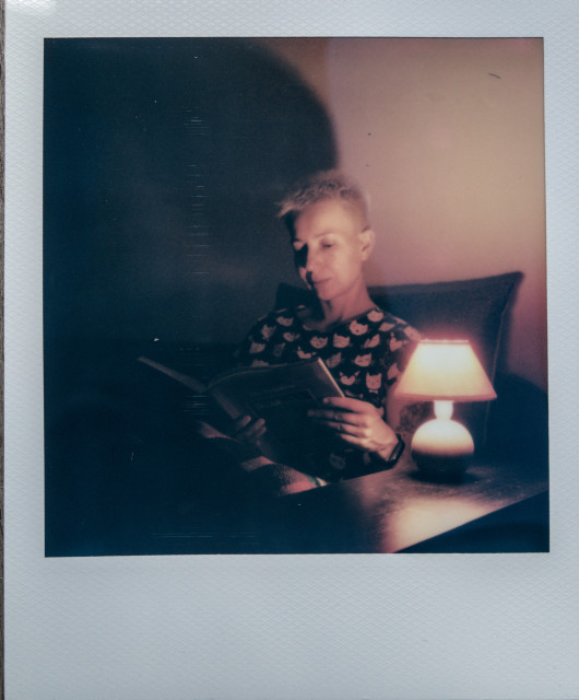 Portrait of a woman sitting on a couch reading a book. A small bedside lamp stands next to her. The lamp illuminates the woman from one side. In general, the image creates an atmosphere of comfort and tranquility