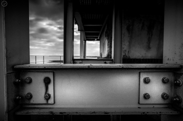 Monochrome shot of girder construction on side of old watch tower. Rusted bolts in details, and cloud covered sky in backdrop