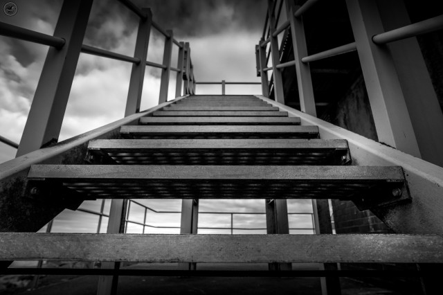 Monochrome shot looking up metal stairs on side of old watch tower, cloud covered sky in backdrop