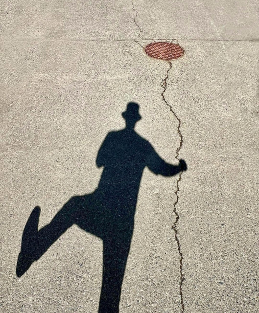 Photography. A color photo of a funny shadow play on a concrete floor. On the one hand, there is the long silhouette of a man in a hat lifting one leg to the side. A long crack runs from the bottom of the picture through the beige concrete floor and extends to a round metal plate at the top of the photo. The whole thing comes together to form a dancing man holding a balloon on a rope. He is about to take off and fly to the sun.