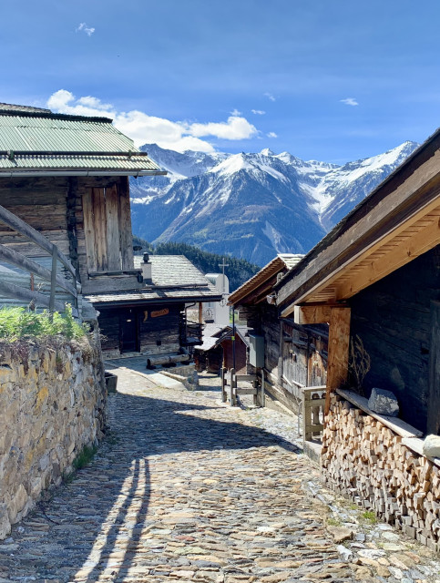 View of a narrow, steep road in a mountain village. To the left and right are old houses made of dark wood. High snow-covered mountains can be seen further back.
