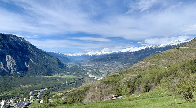 View over a wide valley with spring-like greenery. High mountains to the left and right, a river meanders along the valley below, between a mix of villages, forests and vineyards.
