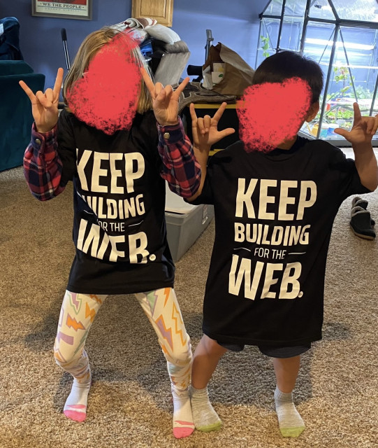 Two kids are wearing Building for the Web black shirts and making gestures with their hands. One is doing heavy metal horns, the other is doing I Love You in sign language.