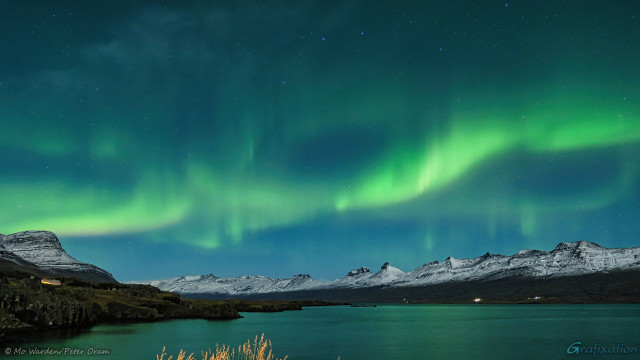 A night photo of a fjord. The mountains surrounding the water on both sides are dark rock capped with snow, lit by moonlight which is coming from behind the viewer. The sky is clear of cloud and many stars are visible, including part of Ursa Major (the Plough or Big Dipper). Across the sky from left to right is a string of green light, some of it showing vertical bar structures, some quite level to the horizontal. Parts of it are brighter than others and, in these places, suggestions of different colours can be seen, yellow fading to a hint of red. The green is so strong that it's reflected in the water surface below.