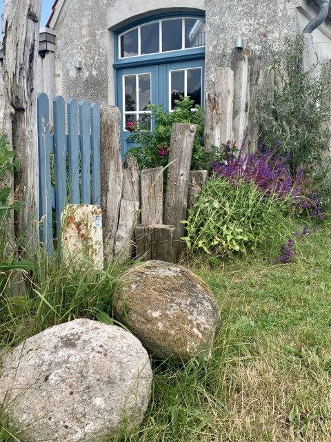 Das Bild zeigt die Front von einem kleinen, alten Haus. Im Vordergrund zwei Findlinge und ein Holzzaun aus alten Balken mit hellblauer Holztür, die Eingangstür hat auch diese Farbe.