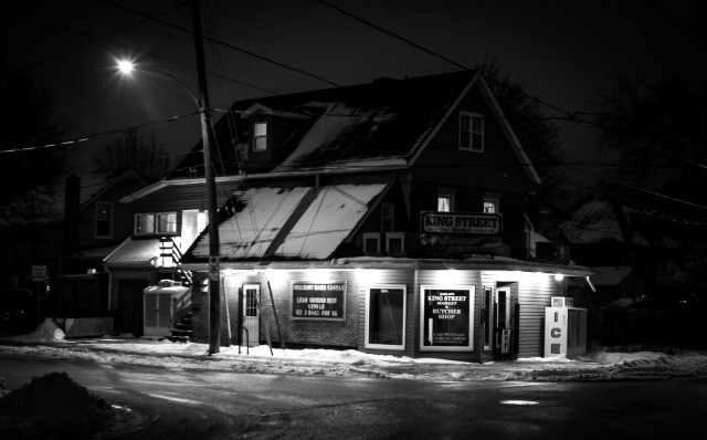 A black and white image of a small brightly lit butcher shop sits on a street corner on a dark winter's night.