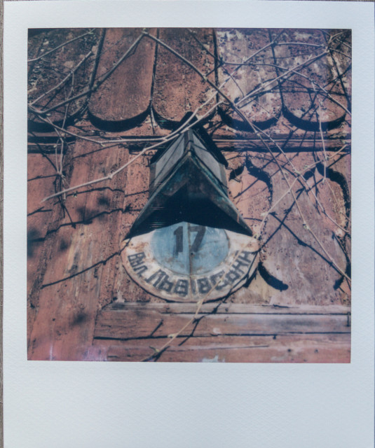 An image of an old house number sign. The sign is metal and has a round shape. It has the house number 17 and the name of the street. Above this sign is a roof in the form of a triangular pyramid. The sign is attached to a wall decorated with carved wooden paneling.