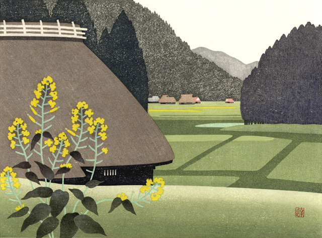 A view of a mountain village in the Japanese countryside, on a spring day. On our left, yellow nanohana flowers screen a view looing down a gently rolling hill toward a large traditional-style thatched roof Japanese farmhouse; beyond, green fields are marked out in a rectangular pattern. Further on, more village houses are scattered along the valley, while tall healthy-looking trees stand close to the fields and march down the side of a nearby mountain. The sky above is overcast, pale and milky white