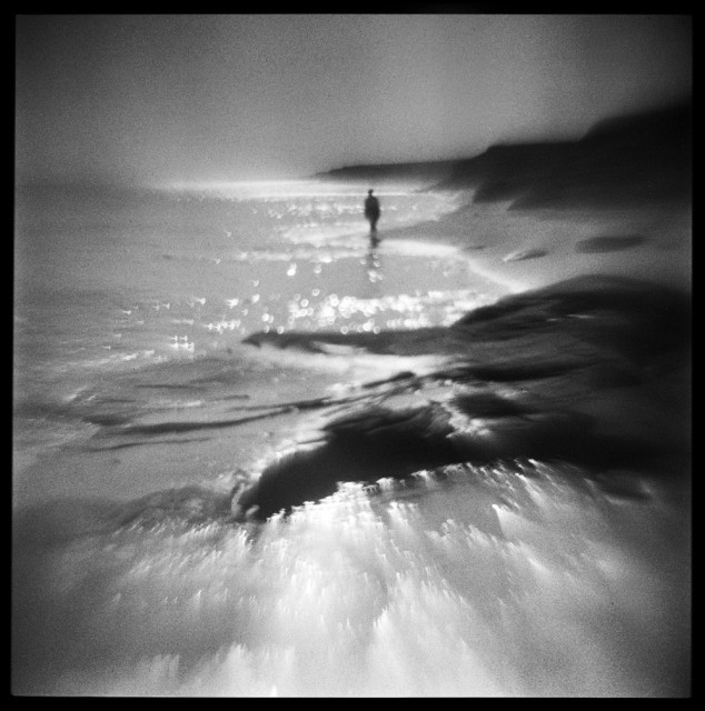 Square black and white image of a figure walking along the ocean shore on the north coast of Prince Edward Island, Canada. Rocks and cliffs line the shore while the morning sun reflects off of the water. The image was created with a modified plastic camera which creates an overall blur to the scene.