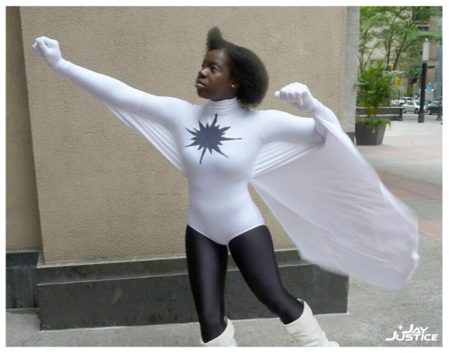 a dark-skinned Black woman with an afro wearing a white leotard with black leggings, and a black starburst on the chest, with long flowing white flight panels under each arm, white gloves, and boots, posed as if about to take flight, with the wind blowing her hair & suit, with a white watermark in the bottom right corner that says Jay Justice