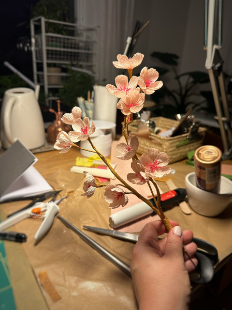 Ett stökigt skrivbord. I fokus en hand som håller en kvist varmrosa blommor i papper.