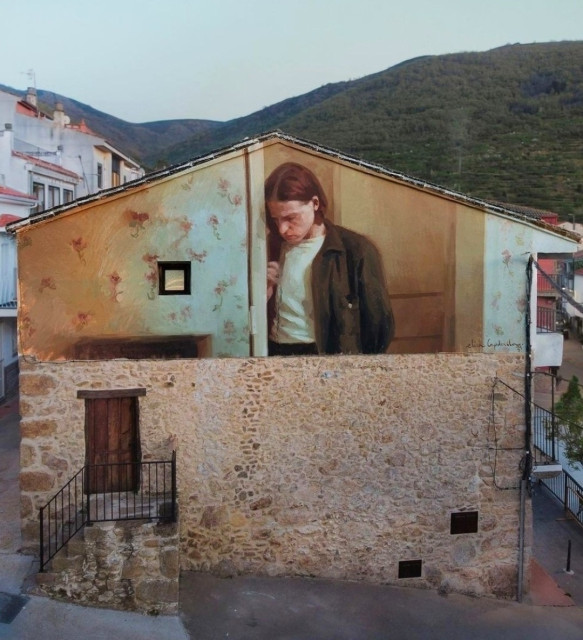 Streetartwall. The mural of a young man has been sprayed/painted on the upper half of the outer wall of a single-storey, old stone building. The young man with medium-length brown hair, white T-shirt and brown-green jacket is standing in front of a painted wooden wall and looking down. A painted wallpaper with a painting and a chest of drawers adorns the left side of the house. It seems like an oversized scene from the house. In the lower part without the mural, the building has a beige-colored stone wall and a staircase with an old wooden door. The photo shows houses in the background and a hilly tree landscape.