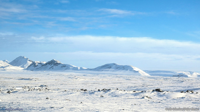 A photo of a snow-covered landscape under a slightly cloudy but blue sky. Sunlight is coming from the right, and catching the faces of a row of mountain ridges in the distance. Some dark, bare rock can be seen at the peaks where the wind has scoured it away. Scarves of light mist are lying just above the ground surface. A road is crossing in front of the viewpoint, and two white vans with side windows are almost certainly carrying tourists to their next beauty spot. It's peaceful, and serene.