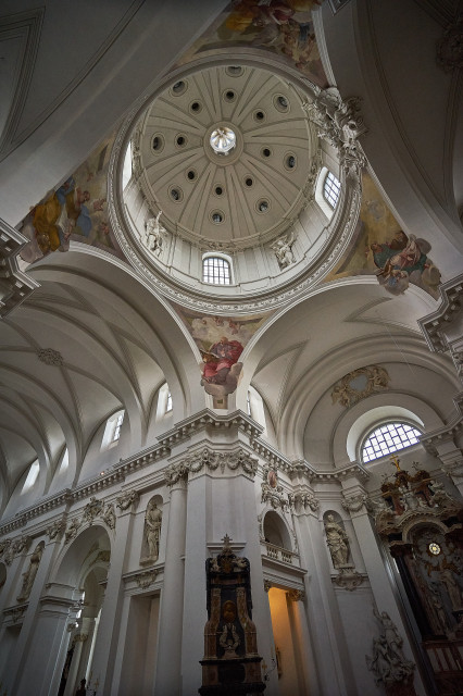 Das Kirchenschiff von Innen. Die große Kuppel an der Decke ist der Mittelpunkt. An der Decke sind Verzierungen in Gold. Von der Mitte gehen die Flügel mit einer Decke aus Kreuzgewölbe weiter.
Die weißen Wände sind mit Marienfiguren verziert.