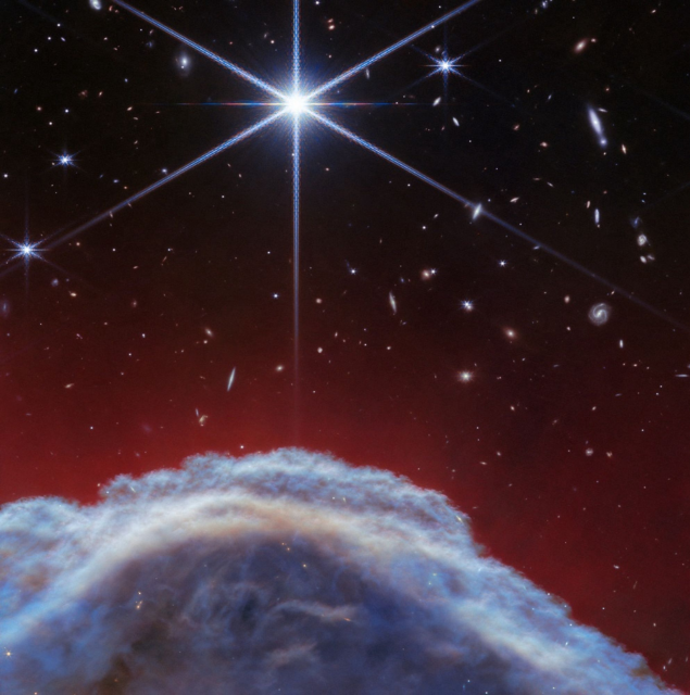 At the bottom of the image a small portion of the Horsehead Nebula is seen close-in, as a curved wall of thick, smoky gas and dust. Above the nebula various distant stars and galaxies can be seen up to the top of the image. One star is very bright and large, with six long diffraction spikes that cross the image. The background fades from a dark red colour above the nebula to black.