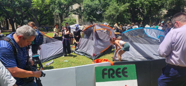 Tents being erected on a campus lawn as signs and people surround them 
