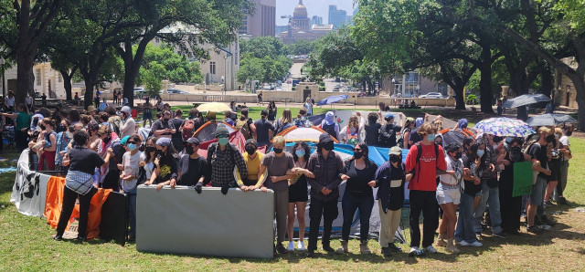 Circular barricades of tables and activists with linked arms protect a group of tents on the campus lawn. 