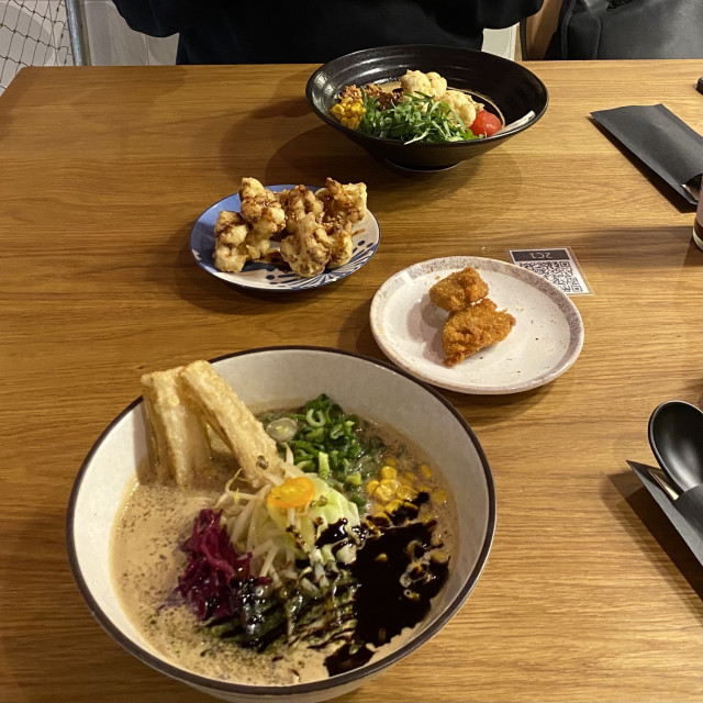 A photo of a table with a white bowl of Ramen in front, a black bowl on the opposite end and two smaller dishes with fried Cauliflower in between. Some cutlery is on the right side of the picture.
The soup in the white dish has a creamy, light color with a large dash of dark liquid, an assortment of veggies, spring onions, corn and some strips that look a little like bacon or krupuk.