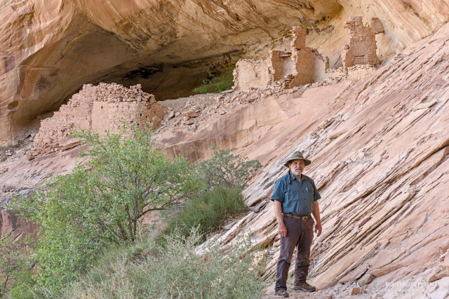 A color photo of a long abandoned cliff-dwelling. The walls have crumbled but you can still tell the dwelling had a dozen rooms and was on two levels. The sandstone cave the dwelling is built in is light tan in color. A small newly leaved out tree is visible in the lower left along with some shrubs. A man stands slightly right of center facing the camera at the bottom of the frame. He has a beard, and wears an olive green narrow brimmed hat, a blue-green short sleeved shirt, dark grey long pants, and boots.