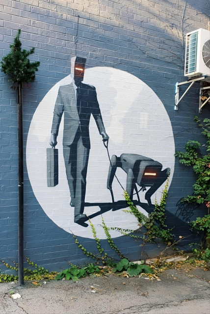 Streetartwall. A mural of a strange figure with a robot head was sprayed/painted on the outside wall of a café. The background is a dark gray brick wall with a light gray circle and implied silhouettes of houses. The figure is wearing a gray business suit, tie, a gray briefcase and has a kind of visor with a glowing slit at eye level instead of a head. He is taking his robot dog for a walk. A futuristic tin dog on thin legs and also with an elongated visor for a head.