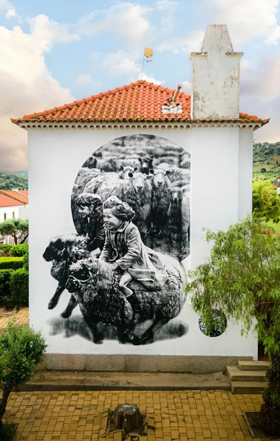 Streetartwall. A black and white mural depicting two children on two sheep was painted on the outside wall of a one-storey old girls' school. The mural is designed like an old photograph. The background is a white wall on which a flock of sheep is depicted in a circle. In front of the circle, two laughing girls in old-fashioned clothes are sitting on two woolly sheep and riding. (The photo shows a long chimney on the right, a red roof, yellow stone slabs and green trees in front of the mural)
Info: The old girls' school has been empty for some time, after a strict separation of girls and boys was no longer desired. This image is based on an original by photographer John Drysdale Title: "Racing on sheep"