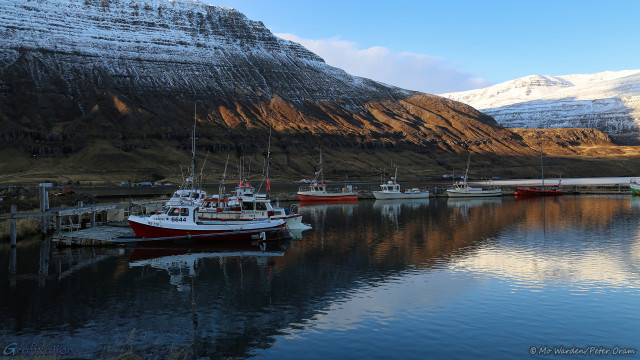 A photo of a dockside comprised mainly of wood. At least six vessels are moored, predominantly small fishing boats. They are mostly white and red painted, with communication aerials and antenna rigged above the cabins. All are floating in a softly rippled harbour, reflecting a craggy mountainside behind the boats. This crinkled brown rock is picking up slanted sunlight from the left, the snow on the heights is still in shadow. Behind this is another white-covered mountain with a mostly blue sky above. It's tranquil and pretty.