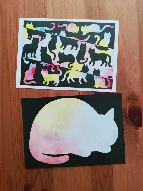 Two postcards on a table, showing the front and back of one design. The front has multiple cats painted in black and colourful gradients. The back is one big cat shape on a black background, that serves as a writing space. 