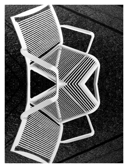 This black and white photograph captures an overhead view of a white, metal, modernist chair positioned on a textured surface that appears to be a patterned floor or paving. The chair's design is characterized by a series of parallel, thin slats that make up the seat and backrest, creating a striking geometric pattern. The positioning of the chair introduces angular shadows that intersect with the chair's structure, adding depth and visual interest to the composition. The contrast between the sleek lines of the chair and the textured ground, along with the play of light and shadow, gives the image a dynamic, abstract quality.