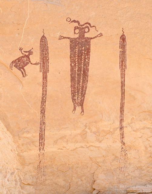A color photo of a pictograph. On the left is a smallish bent over two legged creature with front legs (or arms) spread wide revealing four toes (or figures). The front feet or hands have holes in the palms. Moving to the right is a long rope like image on four long lines surrounded by a pair of shorter lines on either side. All of the lines merge at the top where a short squiggly line emerges as if it were a string to hang the bundle by. This same feature is repeated on the far right, except there are only three center long lines surrounded by single lines on either side. Between the two hanging line figures is a figure with the head of a hammerhead shark, except there are two large hole on each side of the hammer. The body extends down with what appears to be a simple blanket design. There are two short legs with circles at the bottom. And two short arms are spread wide with four fingers each. There's holes in each hand. A snake is above the head.